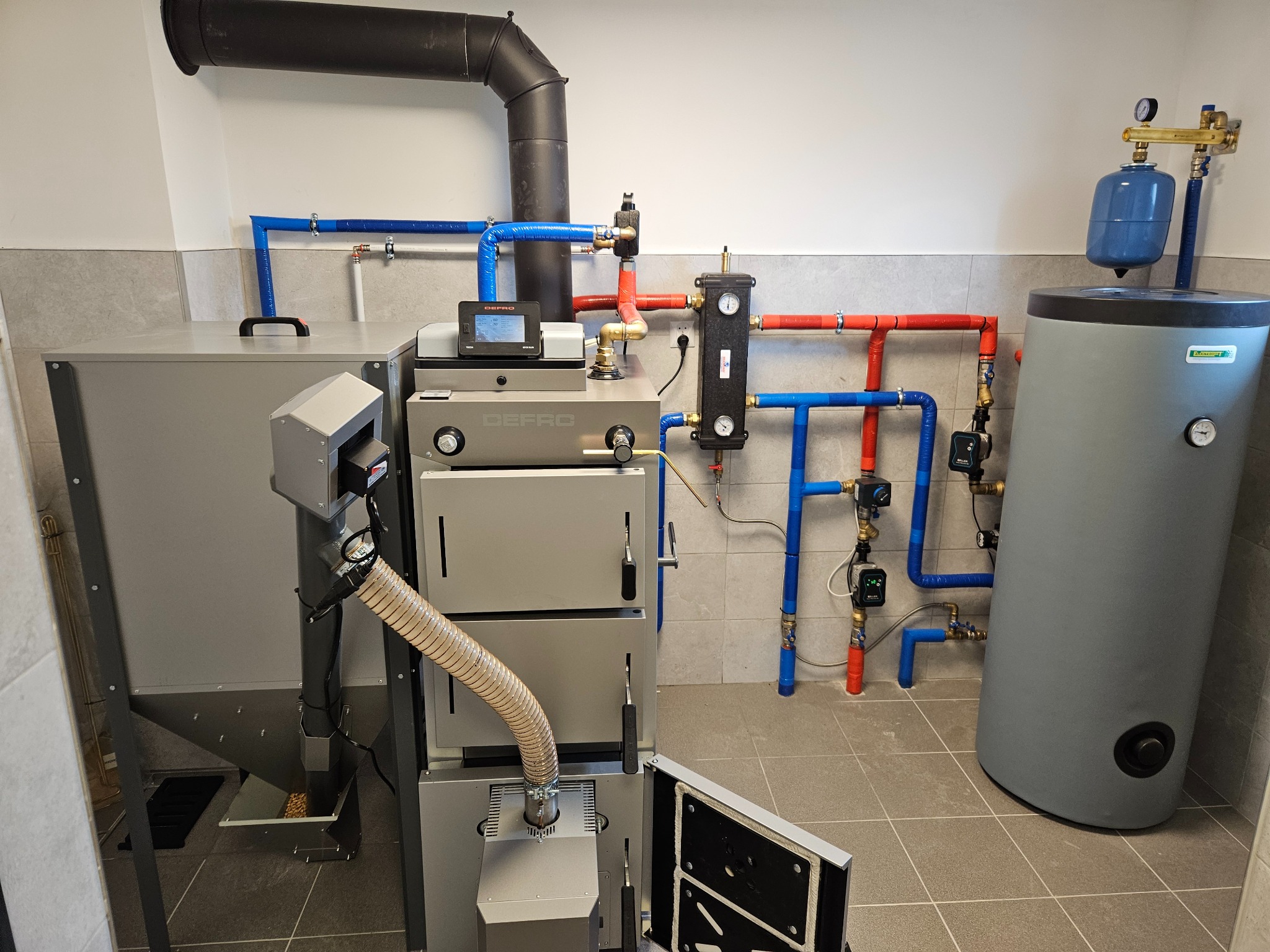 Szary piec CO marki Defro z otwartymi drzwiczkami, widoczne podłączenie rur w kolorach niebieskim i czerwonym, oraz zasobnik na wodę.