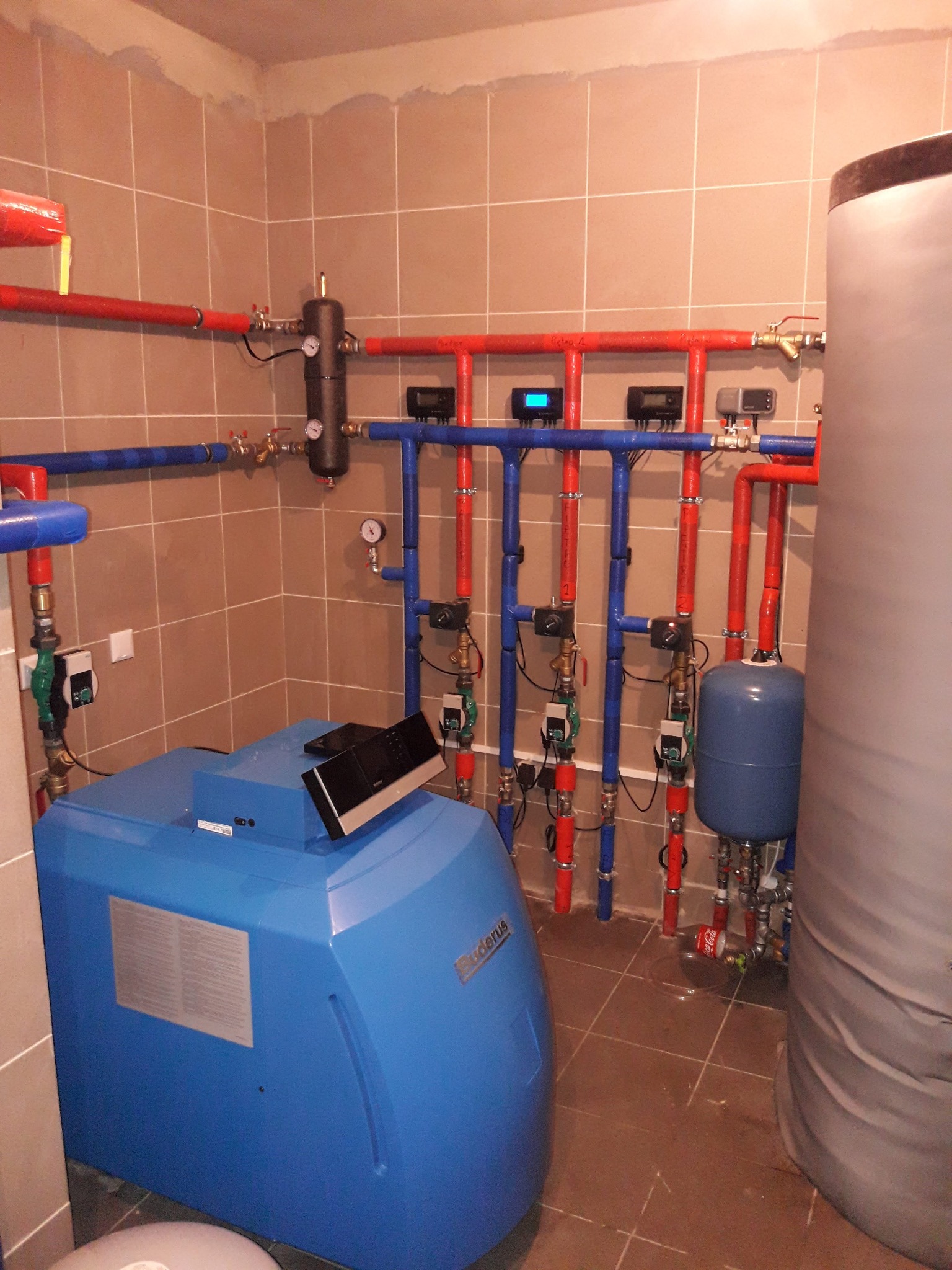 Niebieski piec Buderus z podłączonymi rurami (czerwone i niebieskie) w kotłowni wyłożonej brązowymi płytkami. Widoczny zbiornik akumulacyjny i osprzęt instalacji CO.