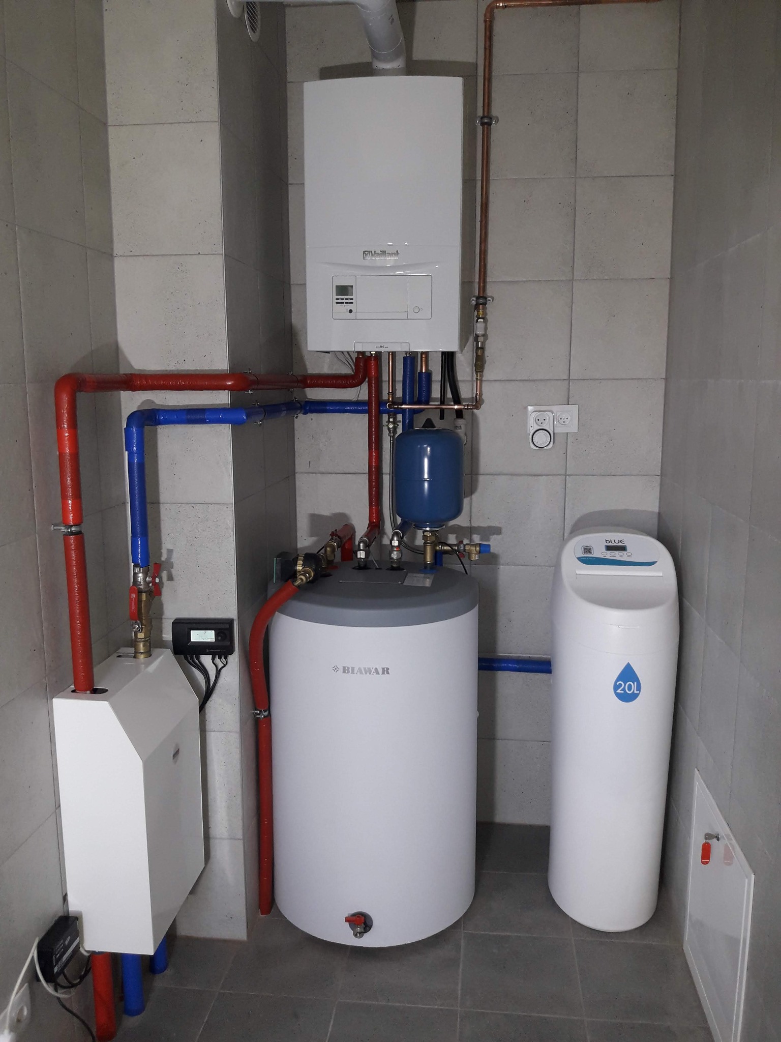 Nowoczesna kotłownia z białym piecem gazowym Vaillant, zasobnikiem Biawar i zmiękczaczem wody Blue 20L na tle szarych płytek. Instalacja w kolorach czerwonym i niebieskim.