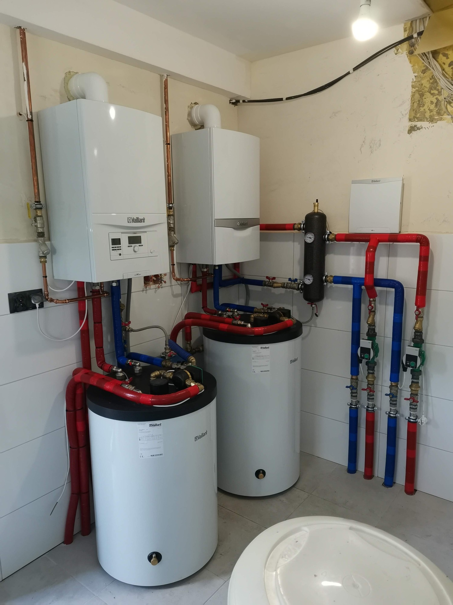 Dwa białe piece gazowe Vaillant z zbiornikami, połączone rurami w kolorach czerwonym i niebieskim, zamontowane w pomieszczeniu z białymi płytkami i fragmentem starej tapety.