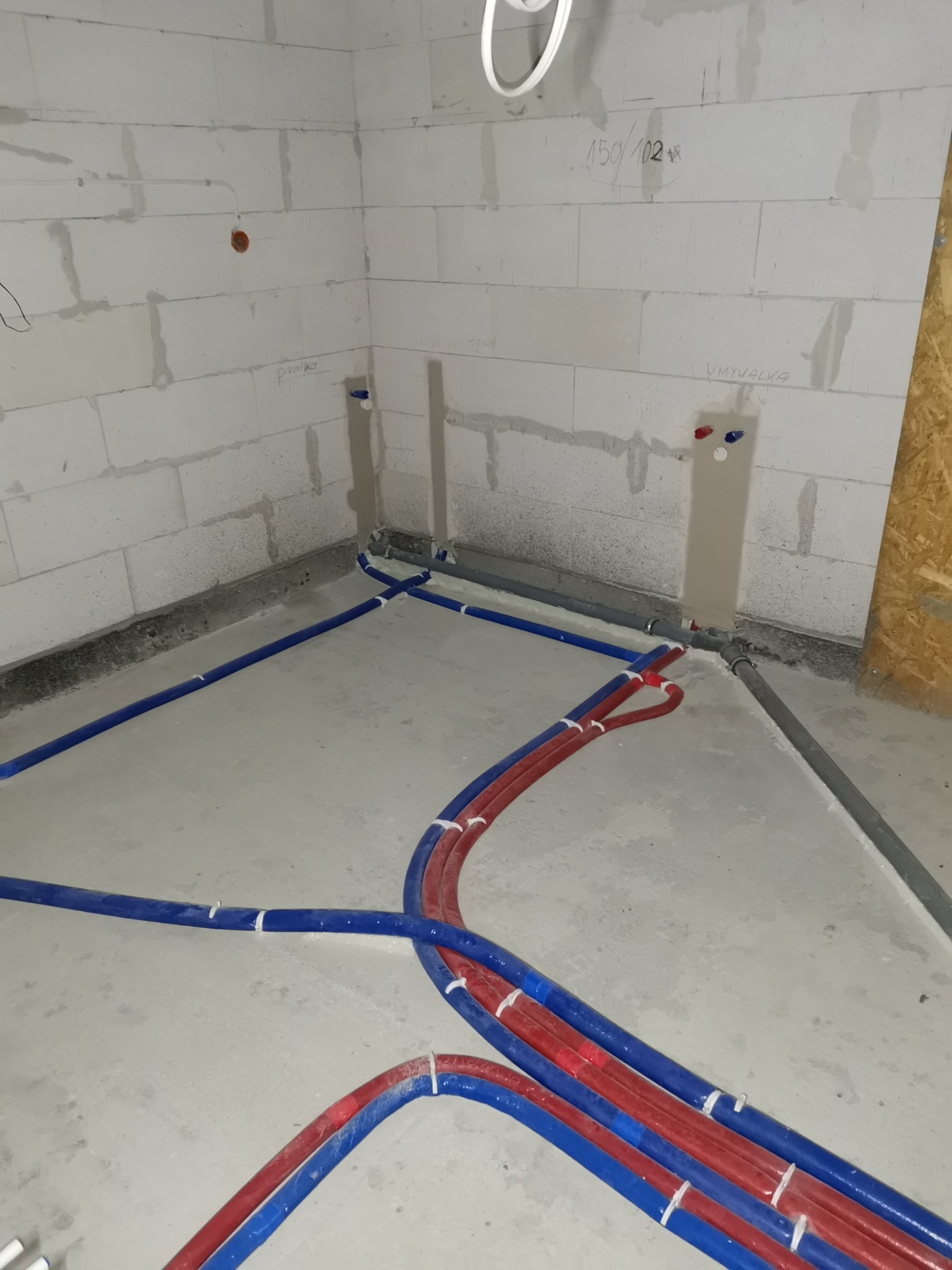 Instalacja wodna w surowym wnętrzu: czerwono-niebieskie rury prowadzone po podłodze, z wyprowadzonymi przyłączami na ścianie z betonu komórkowego.