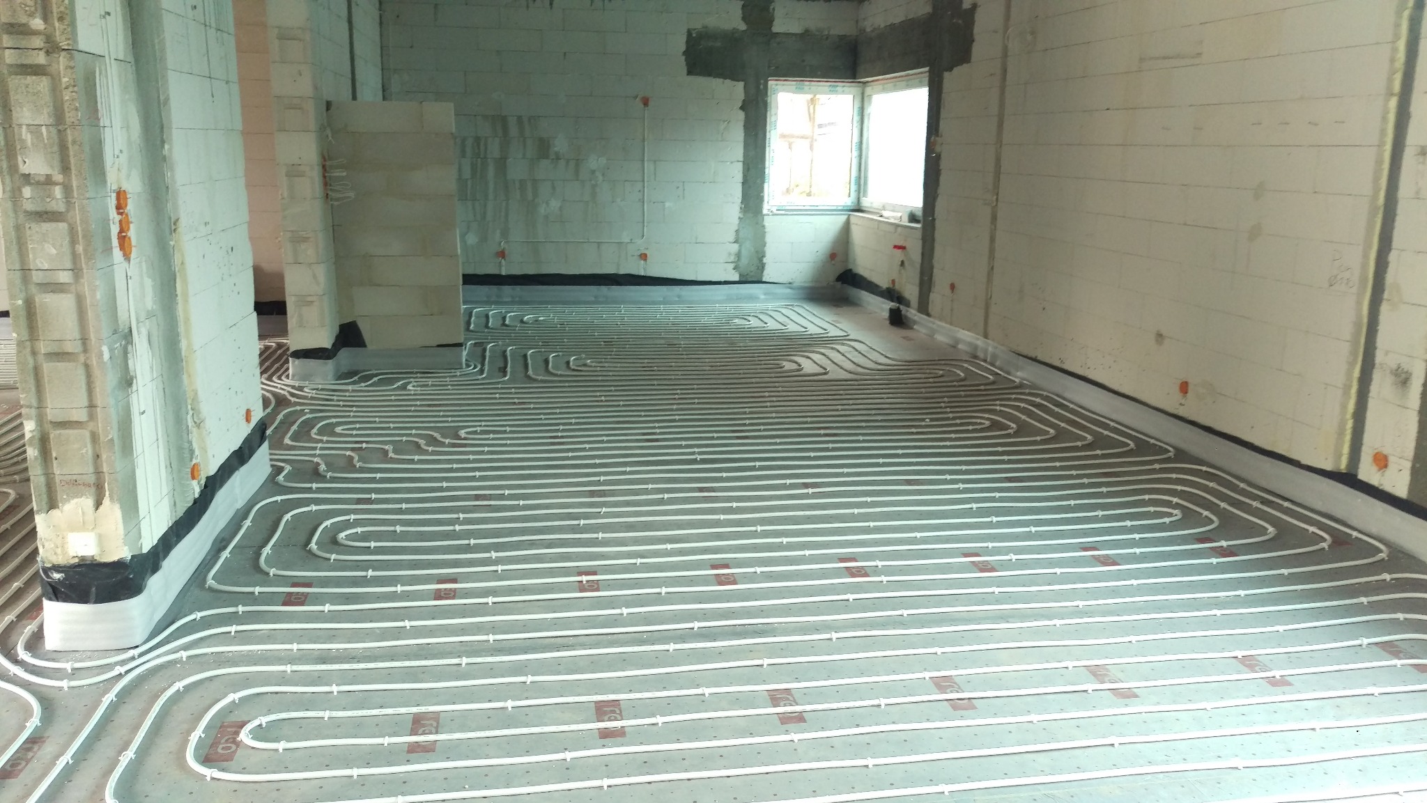 Białe rury ogrzewania podłogowego ułożone w pętle na szarym podłożu w surowym wnętrzu z oknem. Widoczne mocowania rur i izolacja przy ścianach.