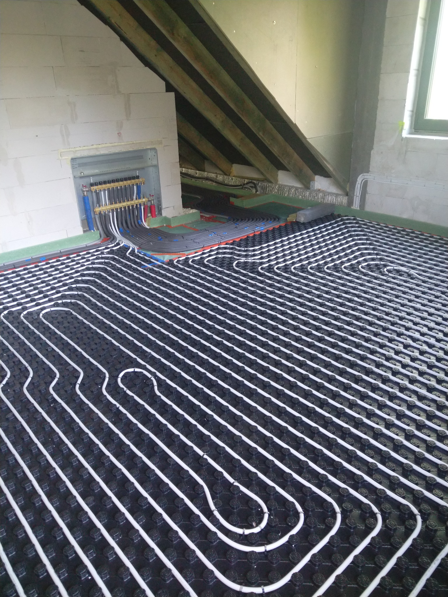 Ogrzewanie podłogowe: białe rury ułożone w pętle na czarnej macie w surowym wnętrzu poddasza, widoczny rozdzielacz i fragment instalacji przy ścianie.