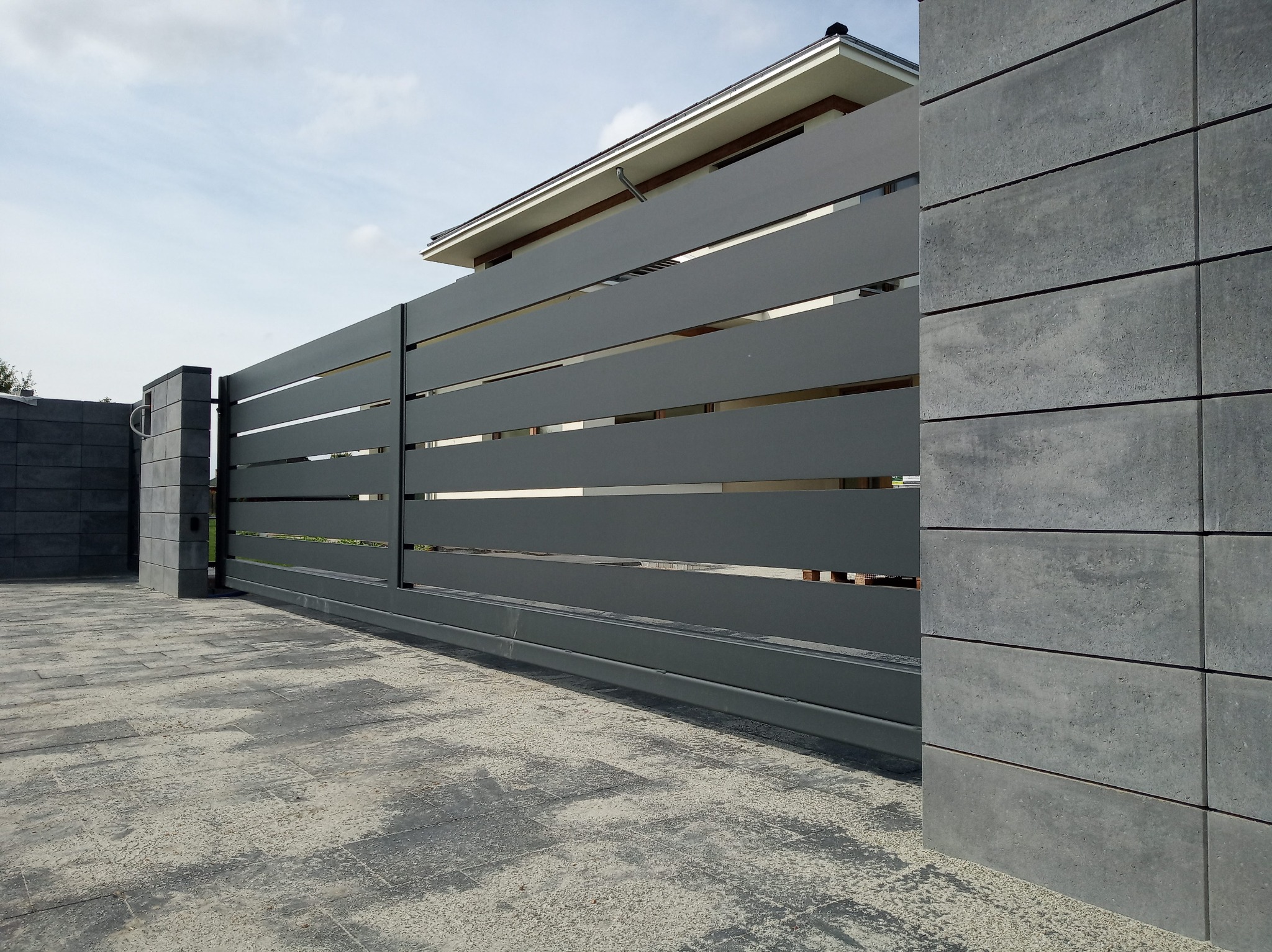 Nowoczesna, przesuwna brama w kolorze grafitowym z poziomymi panelami, zamontowana na posesji z szarą kostką brukową i elementami wykończeniowymi z betonu architektonicznego.