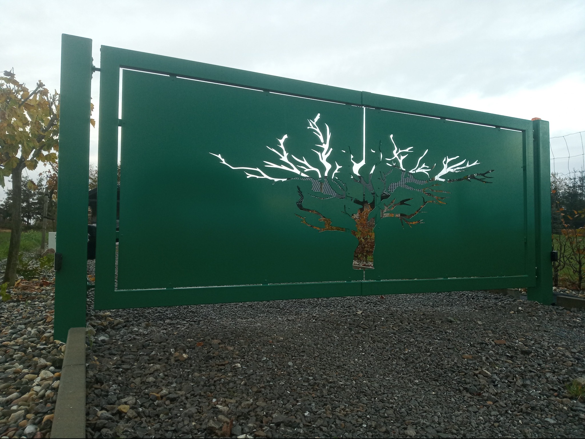 Zielona, metalowa brama z wyciętym motywem drzewa, widoczna na tle pochmurnego nieba i żwirowego podłoża. Minimalistyczny design z precyzyjnymi detalami.
