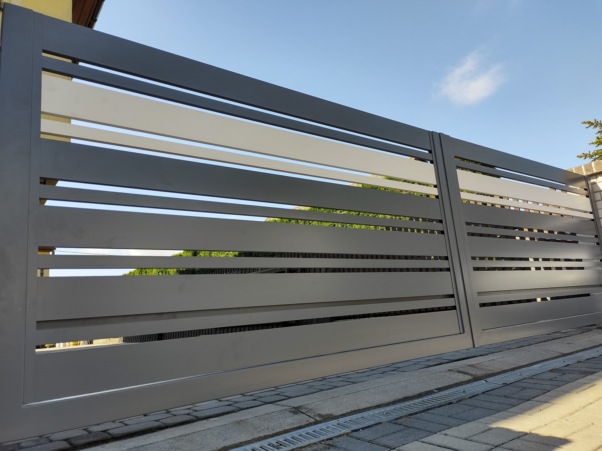 Nowoczesna, przesuwna brama w kolorze szarym z poziomymi panelami, widok z dołu, na tle błękitnego nieba i zieleni. Brama zamontowana na kostce brukowej.