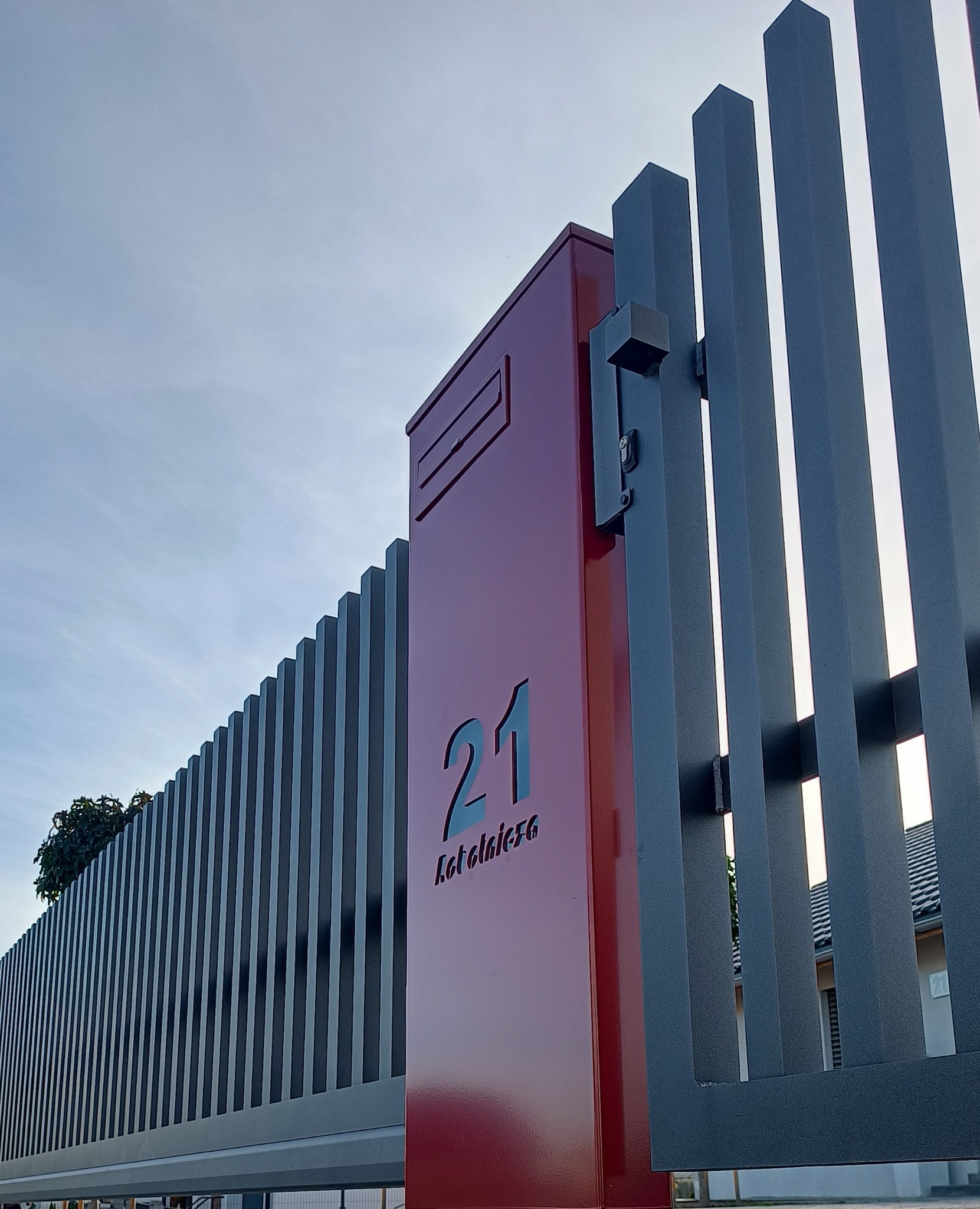 Nowoczesna brama posesyjna z metalowymi sztachetami i skrzynką na listy w Łące. Ciemnoszara konstrukcja z numerem domu wyciętym laserowo, widok z dołu.