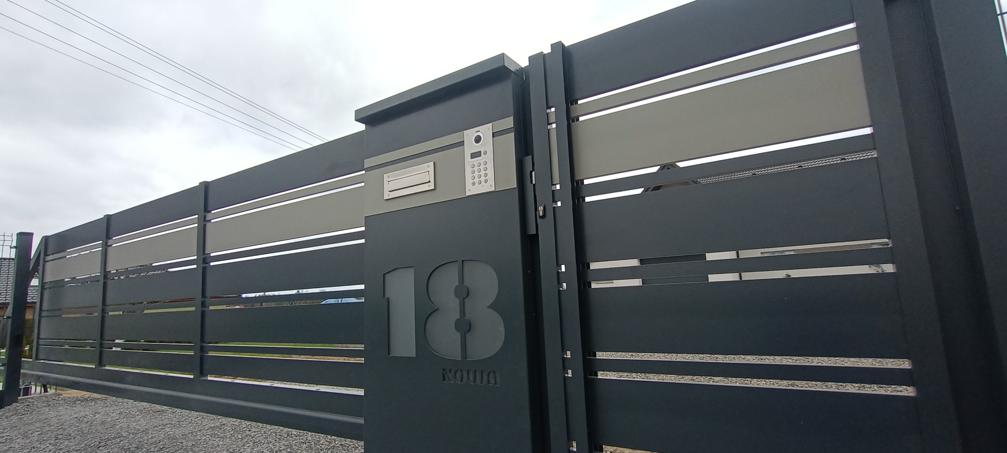 Nowoczesna, przesuwna brama metalowa w kolorze grafitowym z numerem domu '18' i panelem z domofonem, widok z zewnątrz na tle szarego nieba.