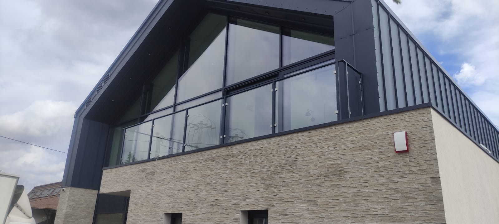 Nowoczesny budynek z balkonem zabezpieczonym szklaną balustradą, wykończony kamieniem elewacyjnym i blachą trapezową na dachu.