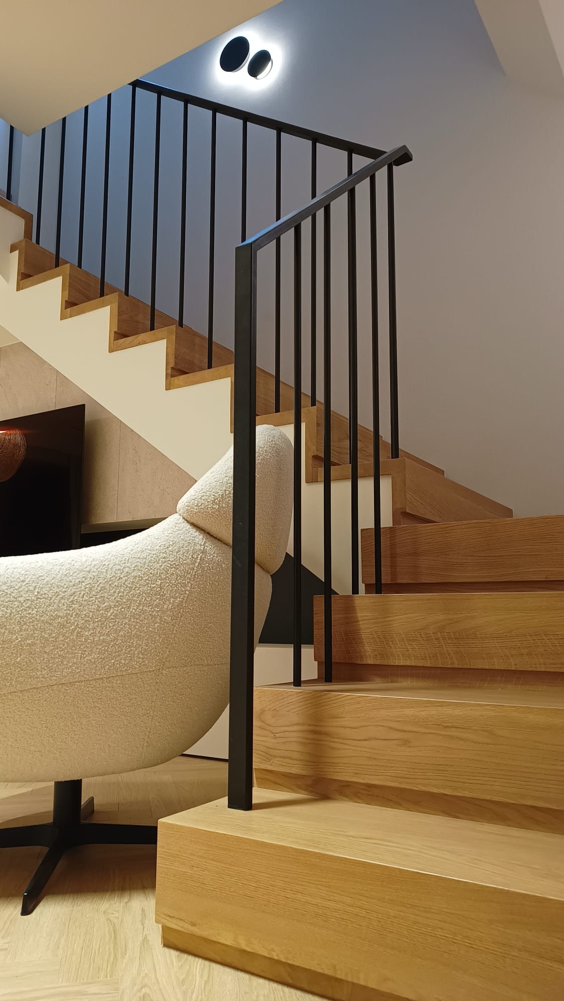Nowoczesne schody drewniane z czarną, metalową balustradą w jasnym wnętrzu. Widoczne stopnie z naturalnego drewna i minimalistyczne oświetlenie sufitowe.