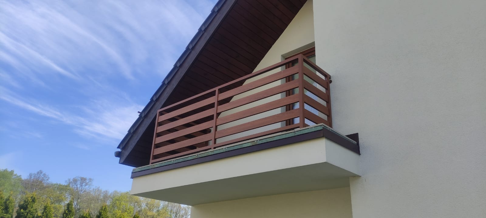 Balkon z drewnianą balustradą w nowoczesnym stylu, widok z dołu na tle błękitnego nieba z chmurami. Uwagę przyciąga kolor balustrady i jej poziomy układ.