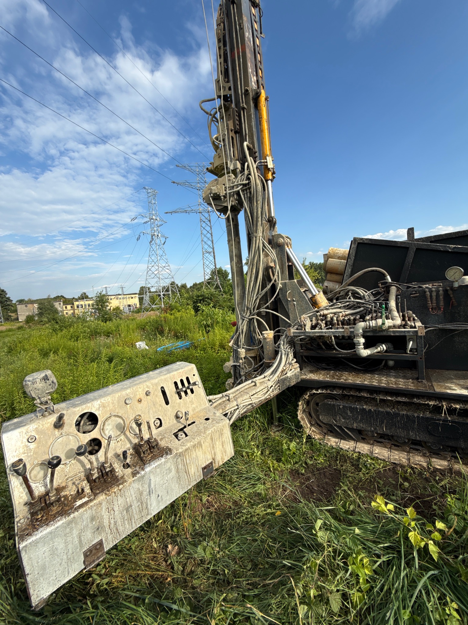Z bliska: Wiertnica na gąsienicach, z widocznym panelem sterowania i wieżą wiertniczą. W tle słupy wysokiego napięcia i budynki. Dzień, błękitne niebo.