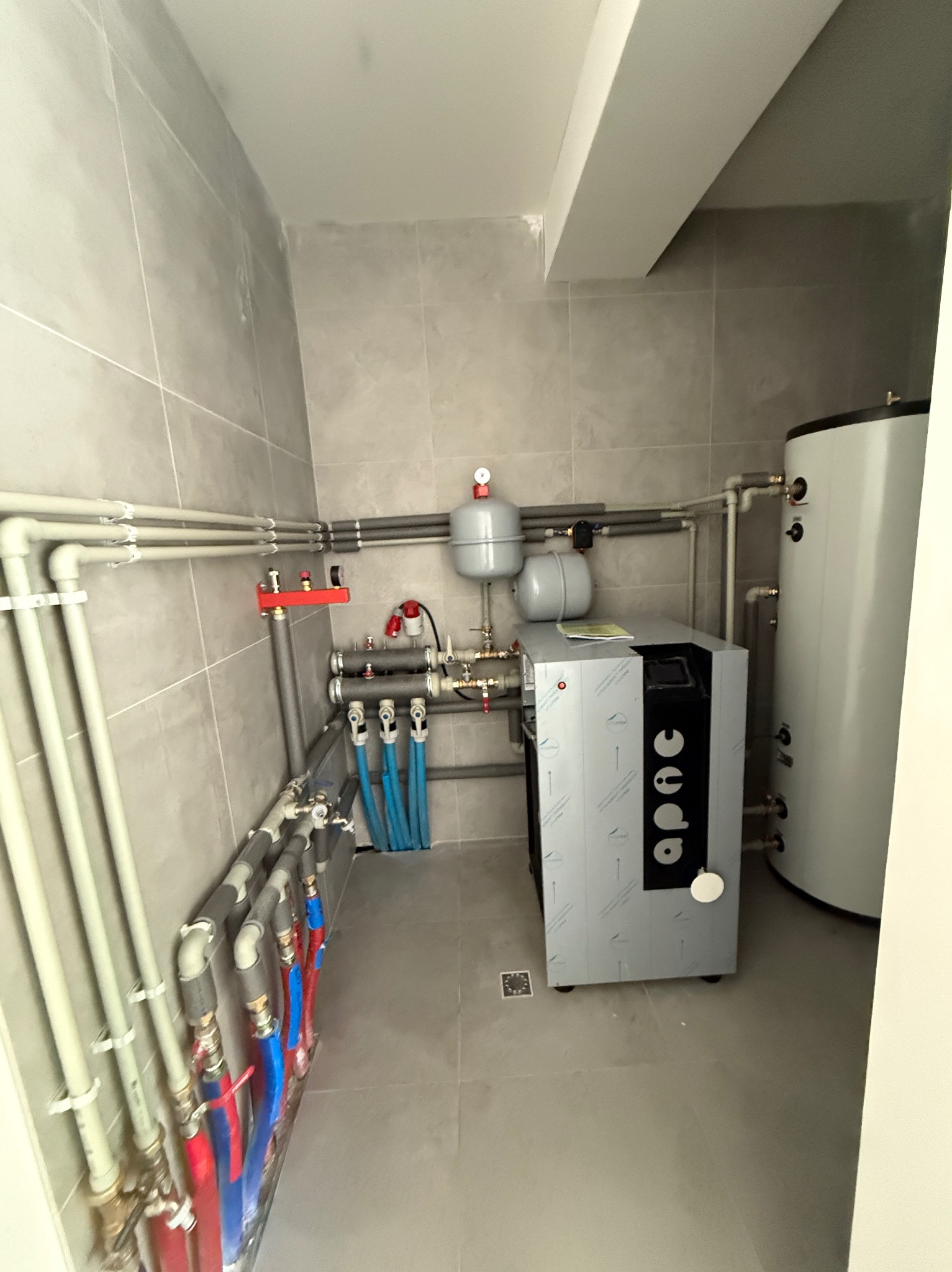 Nowoczesna kotłownia z szarymi płytkami, zbiornikiem i kotłem APAC. Instalacja hydrauliczna z rurami w kolorach szarym, czerwonym i niebieskim.