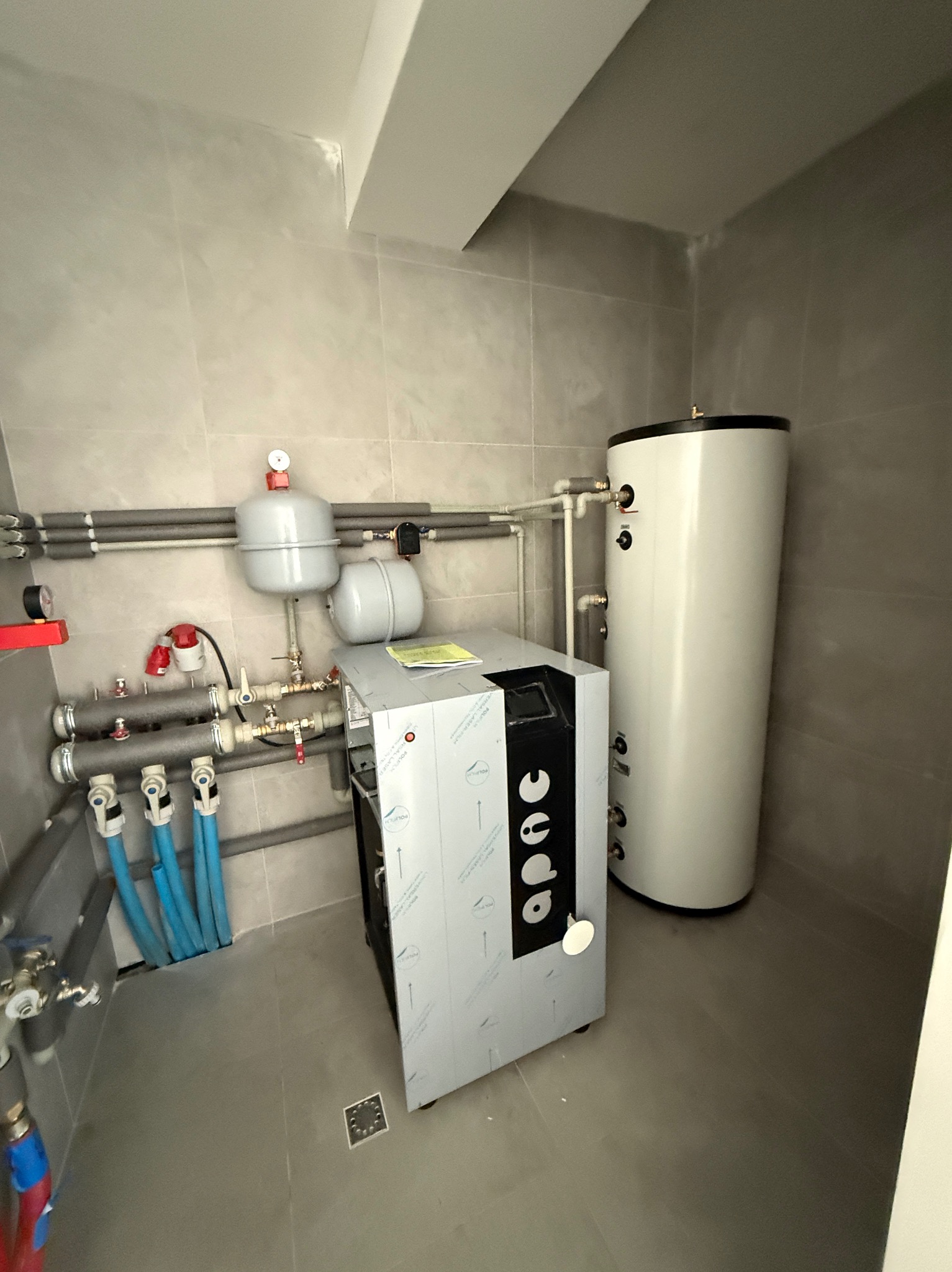 Kotłownia z nowoczesnym piecem APAC, zbiornikiem na wodę i instalacją hydrauliczną na tle szarych płytek. Widoczne rury, zawory i manometry.