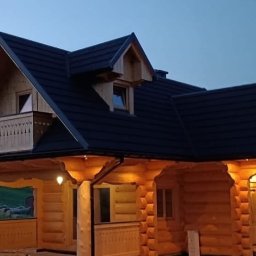 Budownictwo w drewnie Rusnak - Drewniany dom z bali z ciemnym dachem w wieczornym oświetleniu. Widoczny balkon z ozdobną balustradą i oświetlenie zewnętrzne. Charakterystyczna architektura.