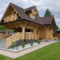 Budownictwo w drewnie Rusnak - Ukończony dom z bali z drewnianym balkonem i schodami wejściowymi wyłożonymi płytkami. Widoczny fragment trawnika i chodnika z kostki.