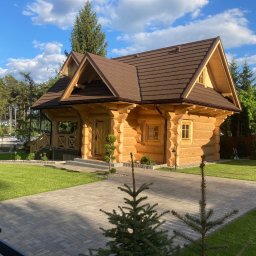 Budownictwo w drewnie Rusnak - Jasny dom z bali z brązowym dachem na tle błękitnego nieba i zieleni. Widoczny podjazd z kostki brukowej i zadbany trawnik. Ujęcie z poziomu gruntu.