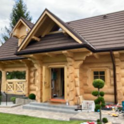 Budownictwo w drewnie Rusnak - Jasny dom z bali z brązowym dachem, widok z zewnątrz. Na pierwszym planie narzędzia i osoba pracująca, co sugeruje trwające prace wykończeniowe.