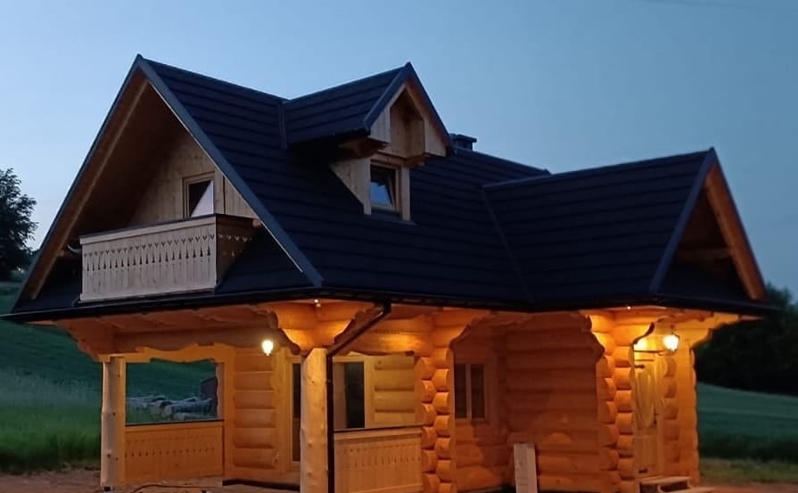 Drewniany dom z bali z ciemnym dachem w wieczornym oświetleniu. Widoczny balkon z ozdobną balustradą i oświetlenie zewnętrzne. Charakterystyczna architektura.