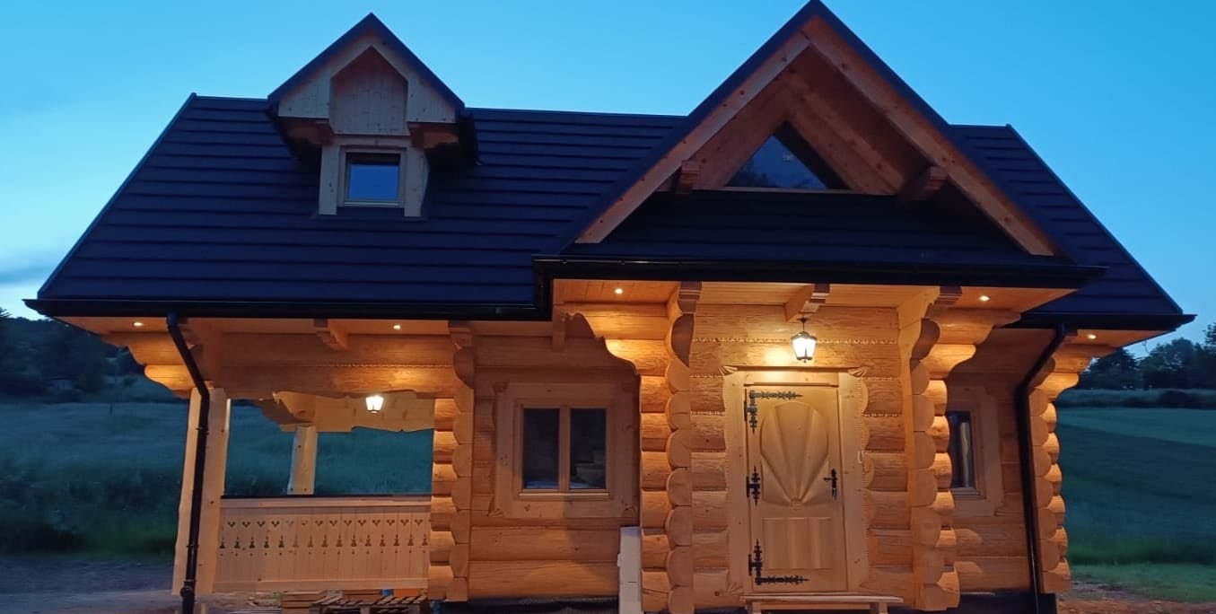 Drewniany dom z bali, oświetlony punktowo, z ciemnym dachem i małym oknem na poddaszu, na tle wieczornego nieba i zieleni.