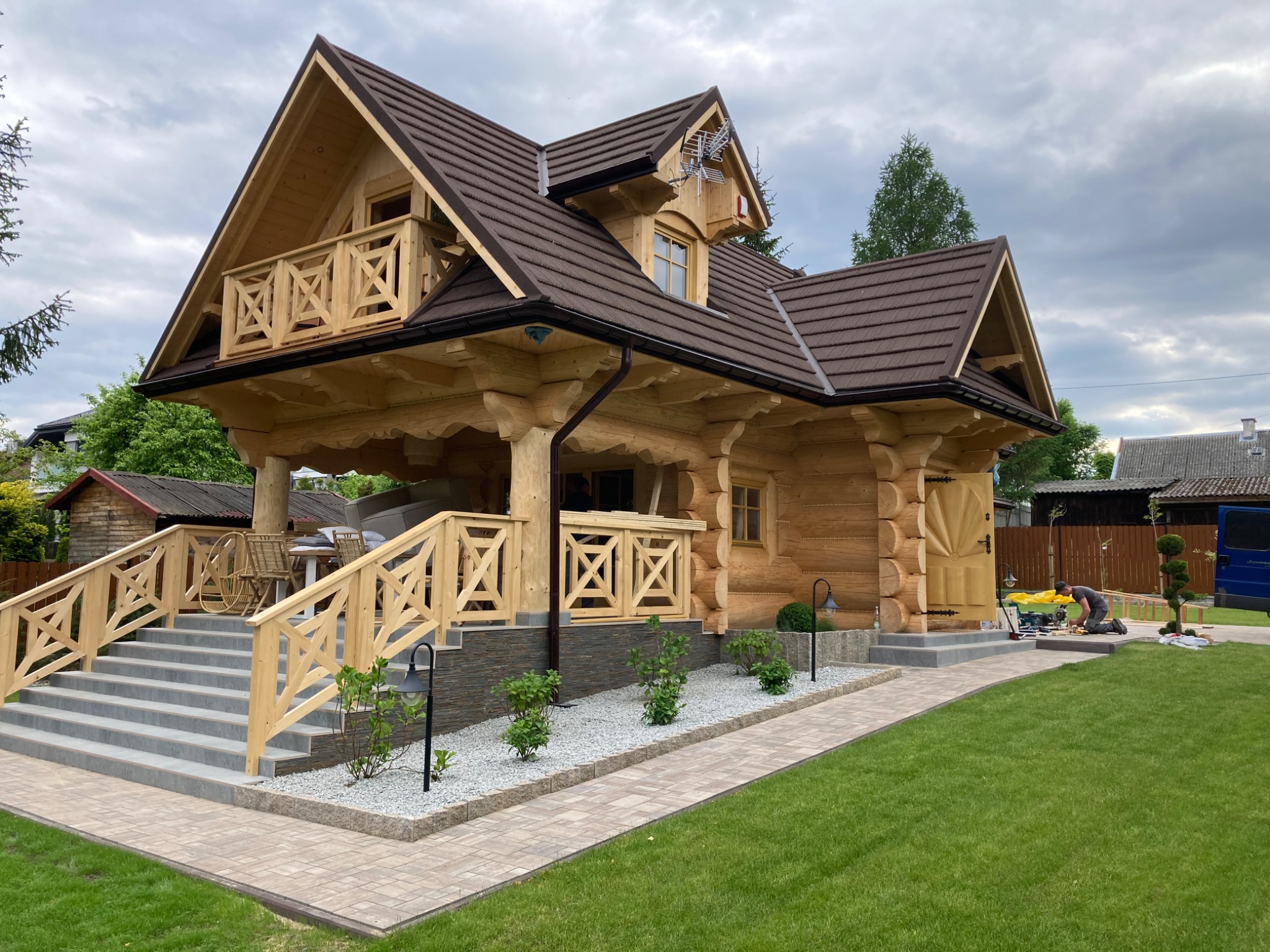 Ukończony dom z bali z drewnianym balkonem i schodami wejściowymi wyłożonymi płytkami. Widoczny fragment trawnika i chodnika z kostki.
