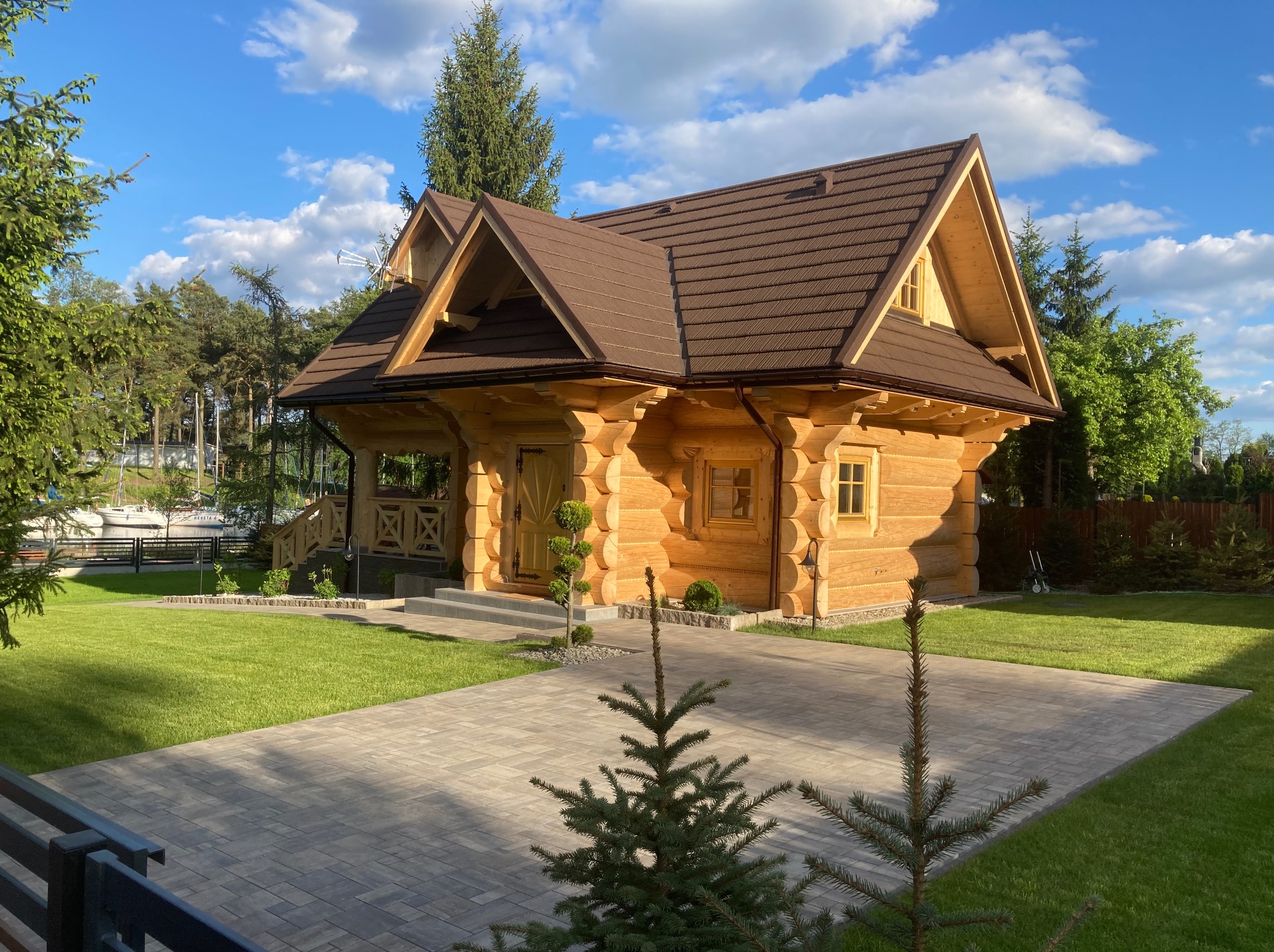 Jasny dom z bali z brązowym dachem na tle błękitnego nieba i zieleni. Widoczny podjazd z kostki brukowej i zadbany trawnik. Ujęcie z poziomu gruntu.