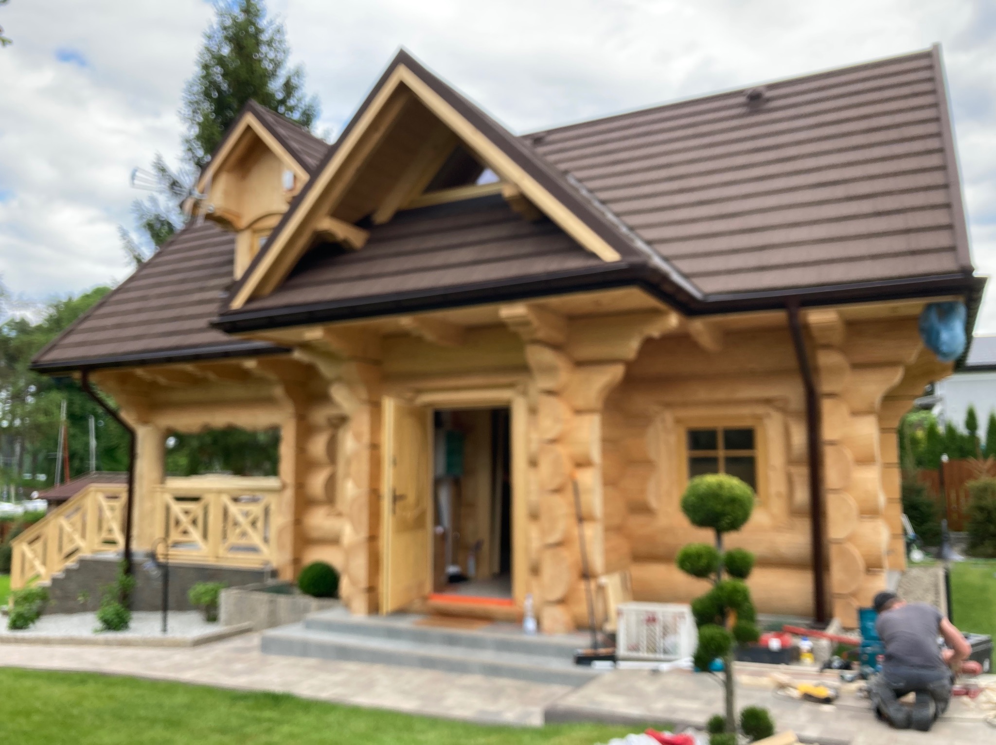 Jasny dom z bali z brązowym dachem, widok z zewnątrz. Na pierwszym planie narzędzia i osoba pracująca, co sugeruje trwające prace wykończeniowe.