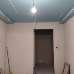 KAMIŃSKI TOMASZ - Pokój w trakcie remontu: zamontowane płyty karton-gips na suficie, ściany przygotowane do malowania, widoczna prowizoryczna żarówka i narzędzia. Otwór drzwiowy bez ościeżnicy.