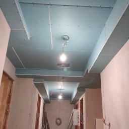 KAMIŃSKI TOMASZ - Korytarz w trakcie montażu sufitu z płyty karton-gips, widoczne profile i oświetlenie. Ściany w stanie surowym, przygotowane do dalszych prac wykończeniowych.