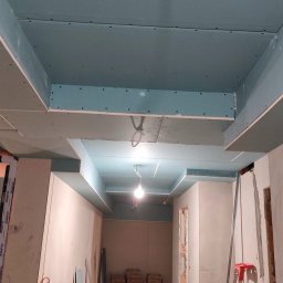 KAMIŃSKI TOMASZ - Sufit podwieszany z płyt karton-gips, widoczne łączenia i otwory na śruby, surowy stan wykończenia, wnętrze w trakcie remontu, drabina oparta o ścianę.