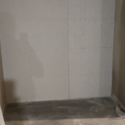 KAMIŃSKI TOMASZ - Ściana z płyt kartonowo-gipsowych w trakcie montażu, widoczne łączenia płyt i instalacja sanitarna w tle. Surowy beton na podłodze, widoczne przewody elektryczne.