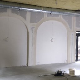 KAMIŃSKI TOMASZ - Wnętrze w trakcie prac wykończeniowych: ściany z łukami po szpachlowaniu, widoczne narzędzia i materiały budowlane, sufit z izolacją.