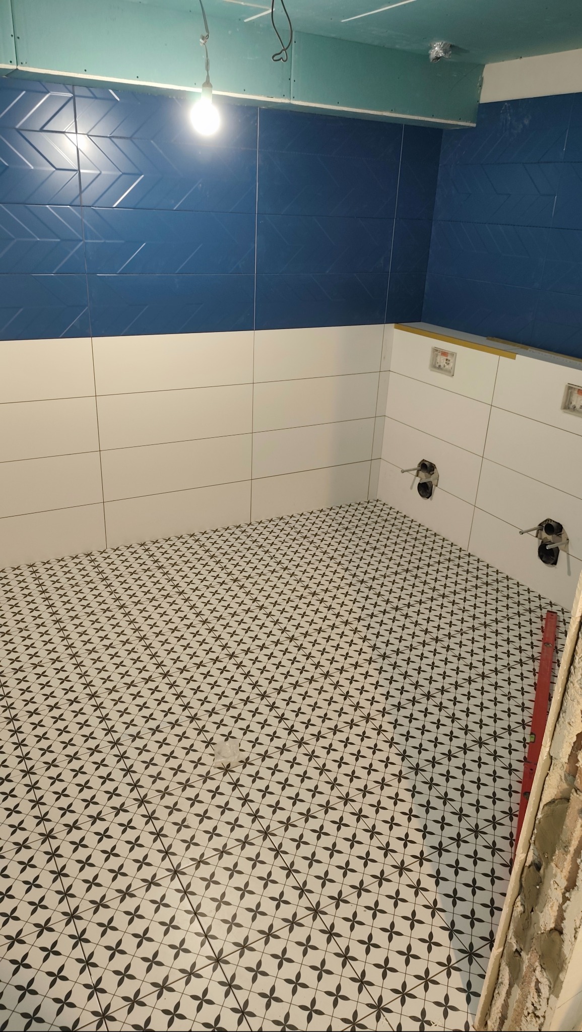 Wykończona łazienka z niebiesko-białymi płytkami na ścianach i wzorzystą podłogą. Widoczne przygotowane punkty instalacyjne. Sufit z płyt kartonowo-gipsowych.