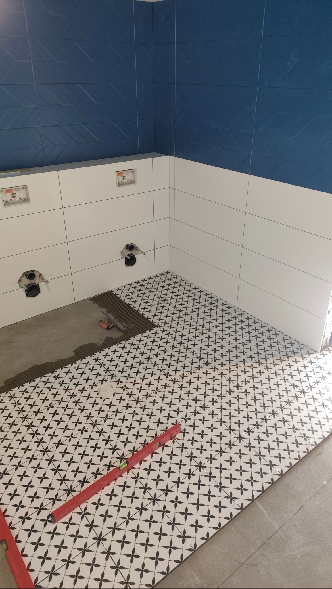 Układanie wzorzystych płytek podłogowych w łazience, częściowo zakończone. Na ścianach białe i niebieskie płytki. Widoczna poziomica i przygotowane otwory na instalacje.