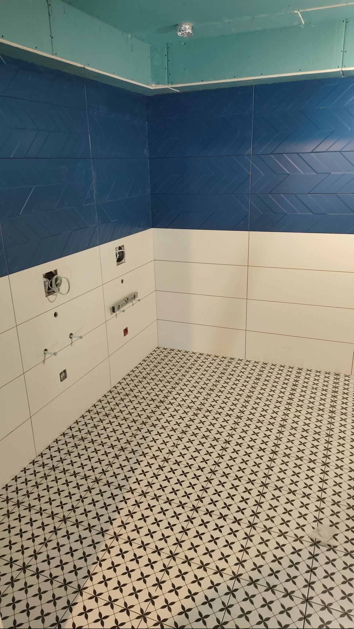 Łazienka w trakcie wykańczania: położone białe i niebieskie płytki ścienne z wzorem oraz czarno-biała mozaika na podłodze. Widoczne instalacje i płyta karton gips na suficie.