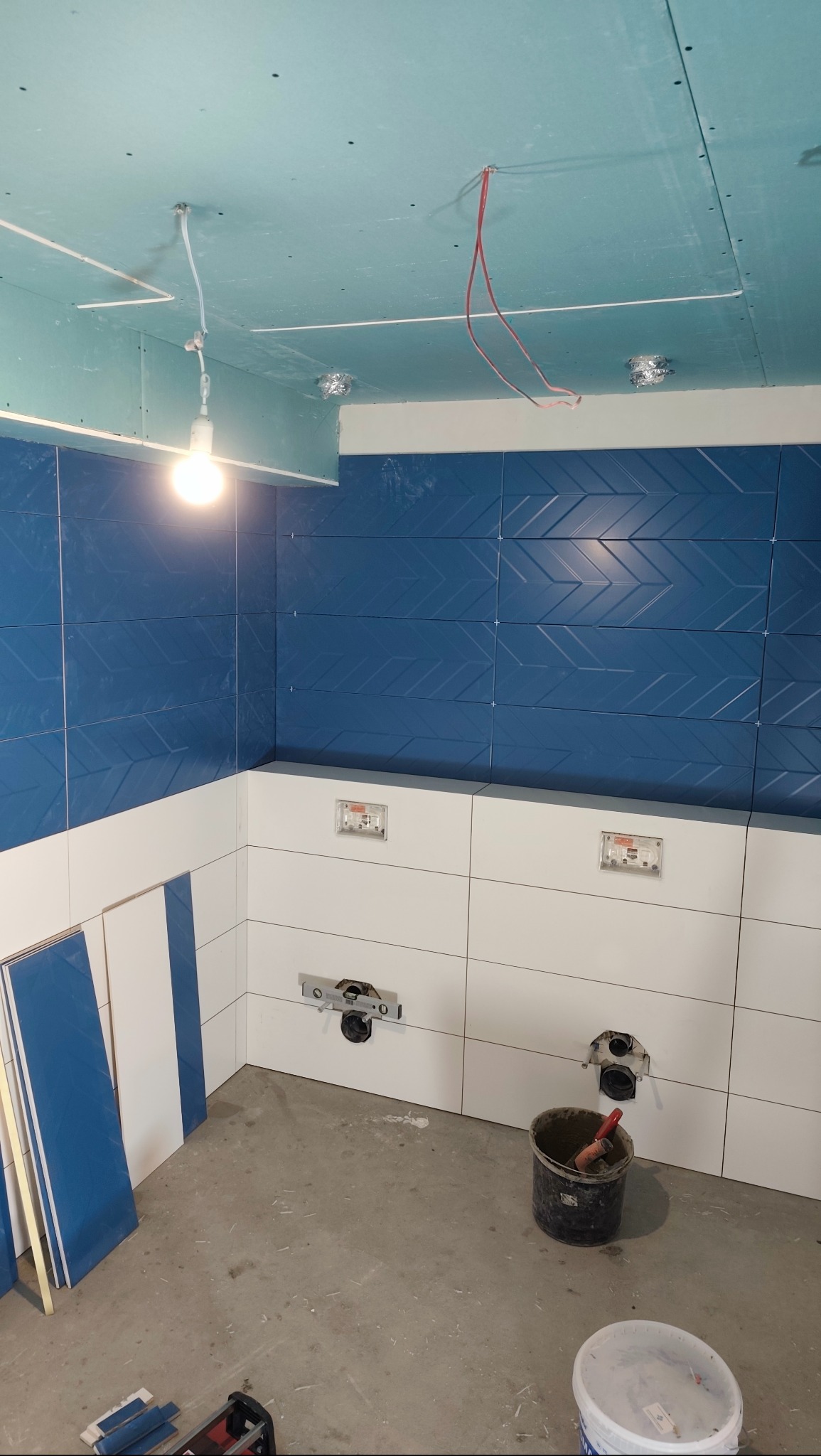 Niebiesko-biała łazienka w trakcie wykańczania: ułożone płytki, widoczne instalacje, sufit z płyt karton-gips, narzędzia glazurnicze. Wnętrze w surowym stanie.