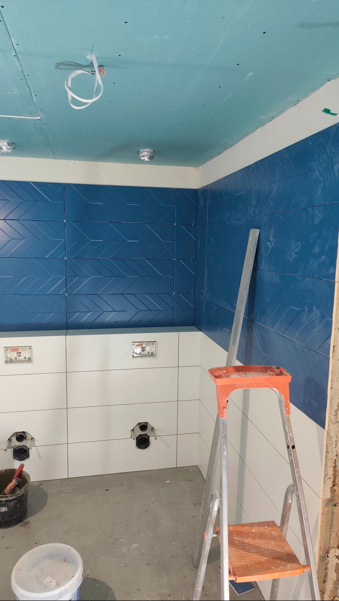 Łazienka w trakcie wykańczania: niebieskie płytki z wzorem jodełki na ścianie, białe płytki poniżej, widoczne stelaże pod WC, drabina i sufit z płyt kartonowo-gipsowych.