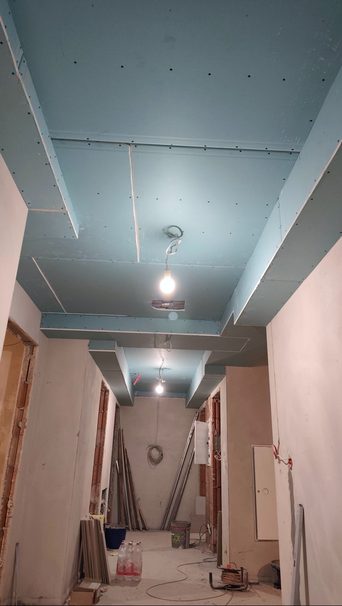 Korytarz w trakcie montażu sufitu z płyty karton-gips, widoczne profile i oświetlenie. Ściany w stanie surowym, przygotowane do dalszych prac wykończeniowych.