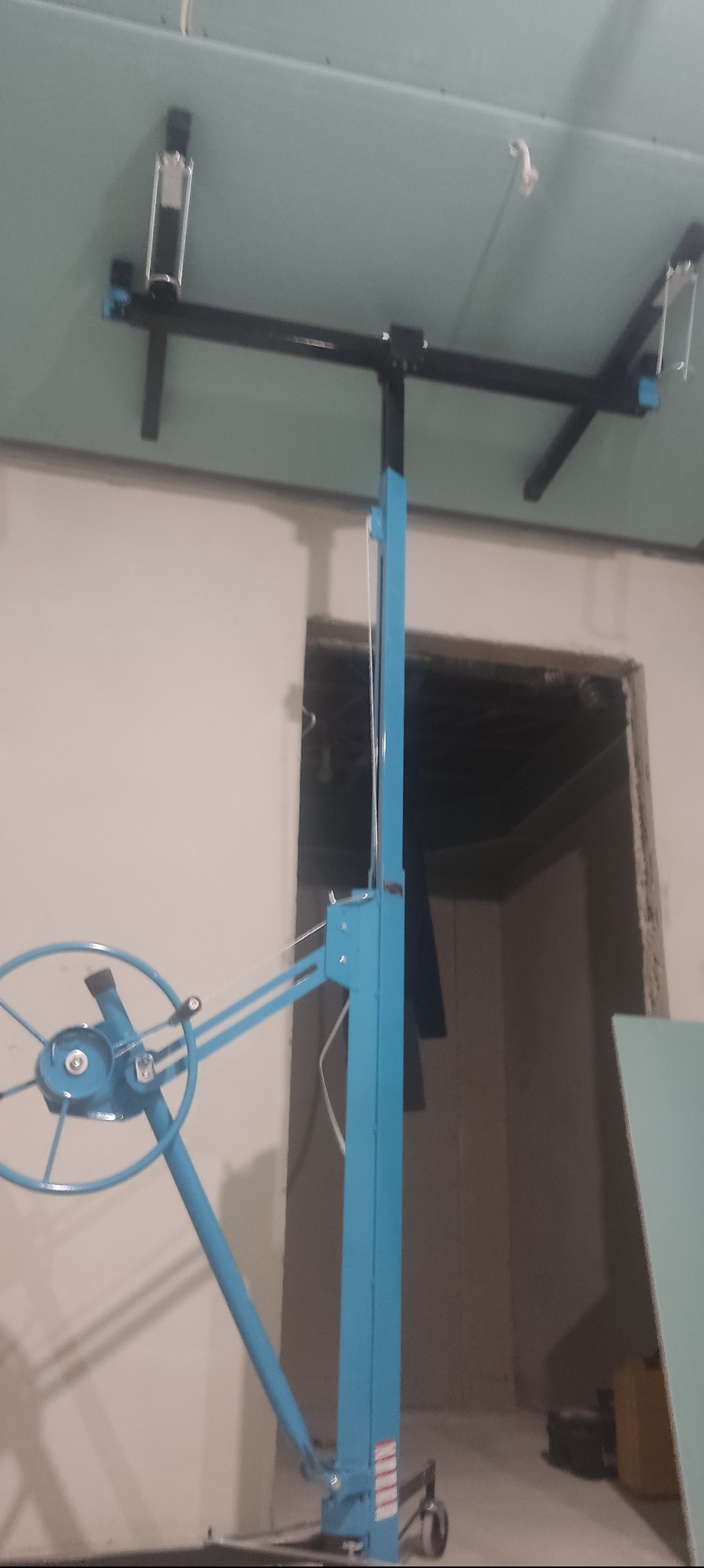 Niebieski podnośnik do płyt gipsowych podtrzymuje zieloną płytę przy suficie. W tle widoczne surowe ściany i otwór drzwiowy. Etap prac wykończeniowych.