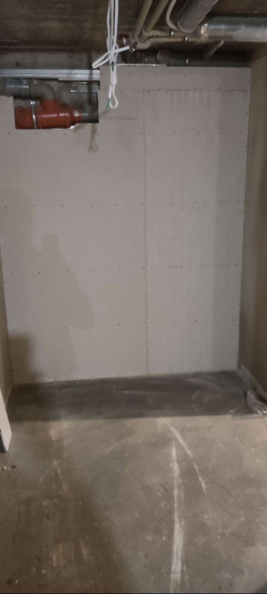 Ściana z płyt kartonowo-gipsowych w trakcie montażu, widoczne łączenia płyt i instalacja sanitarna w tle. Surowy beton na podłodze, widoczne przewody elektryczne.
