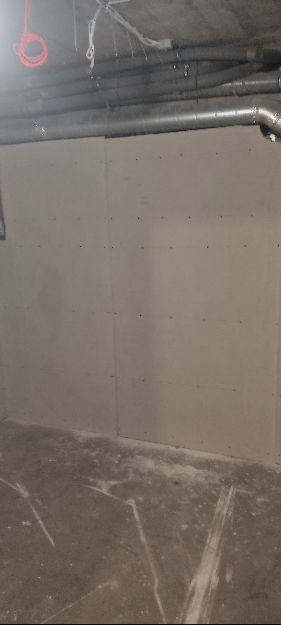 Ściana z płyt kartonowo-gipsowych, widoczne łączenia i śruby, surowy beton na podłodze, instalacje pod sufitem. Wnętrze w trakcie prac wykończeniowych.