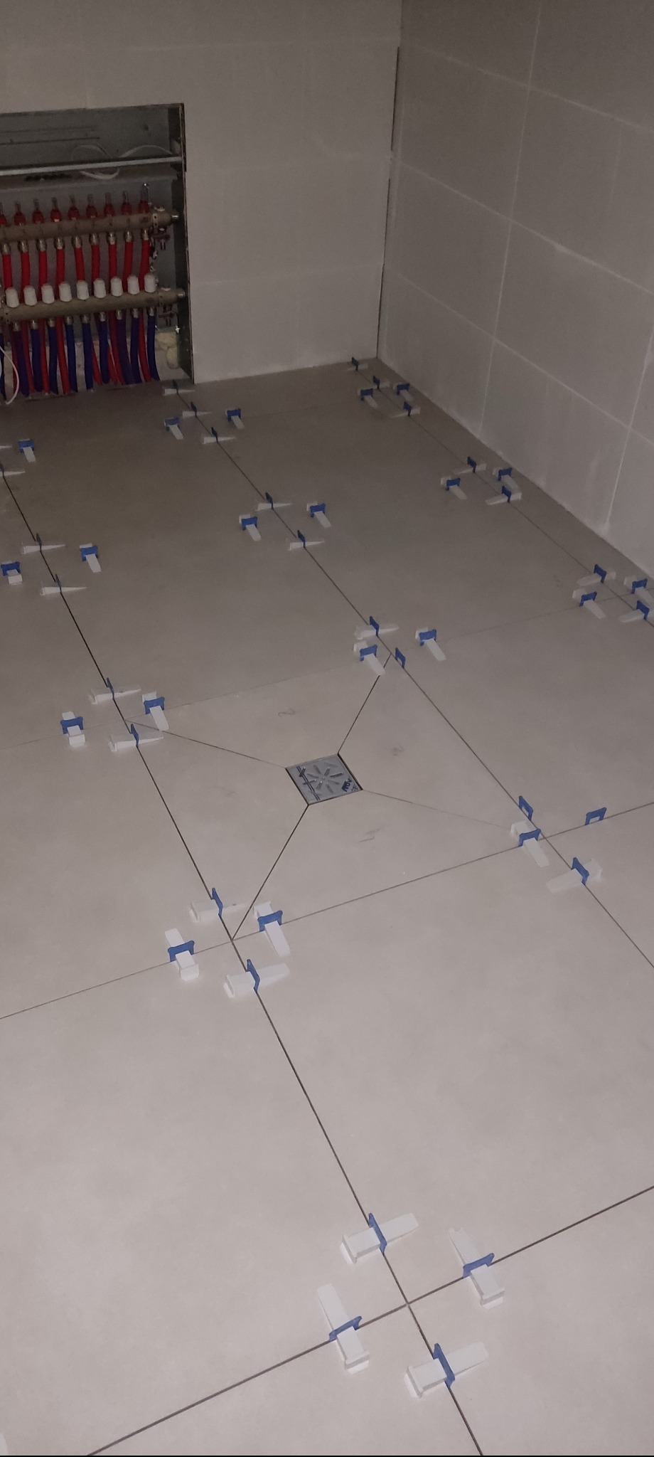 Układanie jasnych płytek podłogowych w łazience z użyciem systemu poziomowania. Widoczny odpływ liniowy i instalacja ogrzewania podłogowego w ścianie.