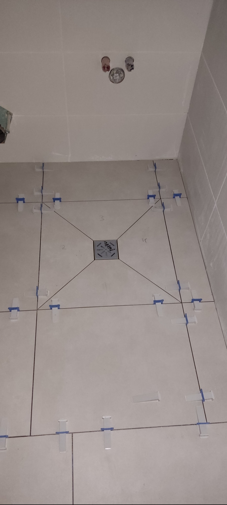 Układanie płytek podłogowych w łazience z użyciem systemu poziomowania. Widoczny odpływ liniowy i krzyżyki dystansowe. Etap prac przed fugowaniem.