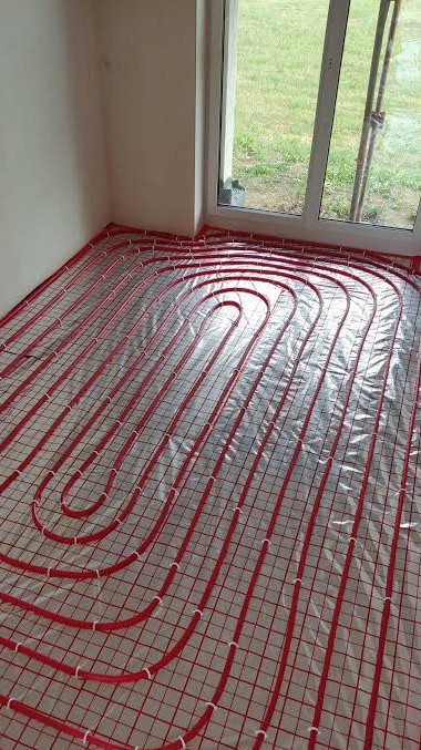 Czerwone rury ogrzewania podłogowego ułożone na folii, widoczne białe uchwyty mocujące, w tle okno z widokiem na zieleń.