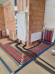 Instalacja ogrzewania podłogowego w trakcie realizacji: czerwono-niebieskie rury ułożone na podłodze, ściany z cegły, piec zabezpieczony folią. Widok z góry.