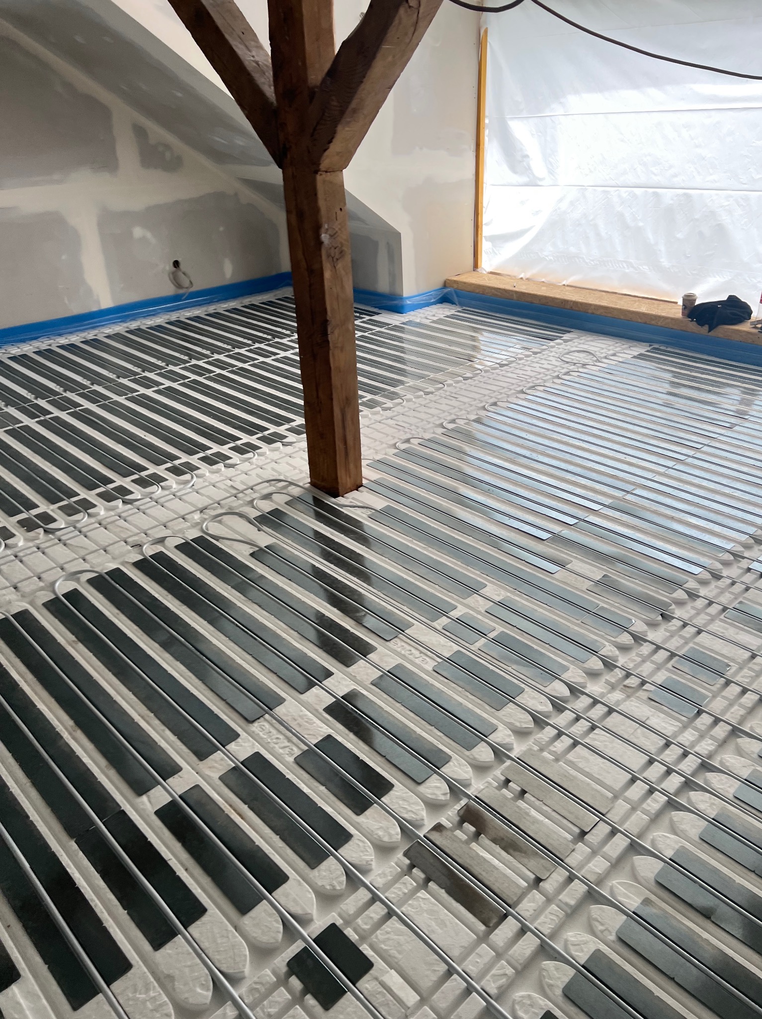 Instalacja ogrzewania podłogowego w poddaszu z widoczną drewnianą belką konstrukcyjną. System ułożony na białej macie, zabezpieczony niebieską folią przy ścianach.