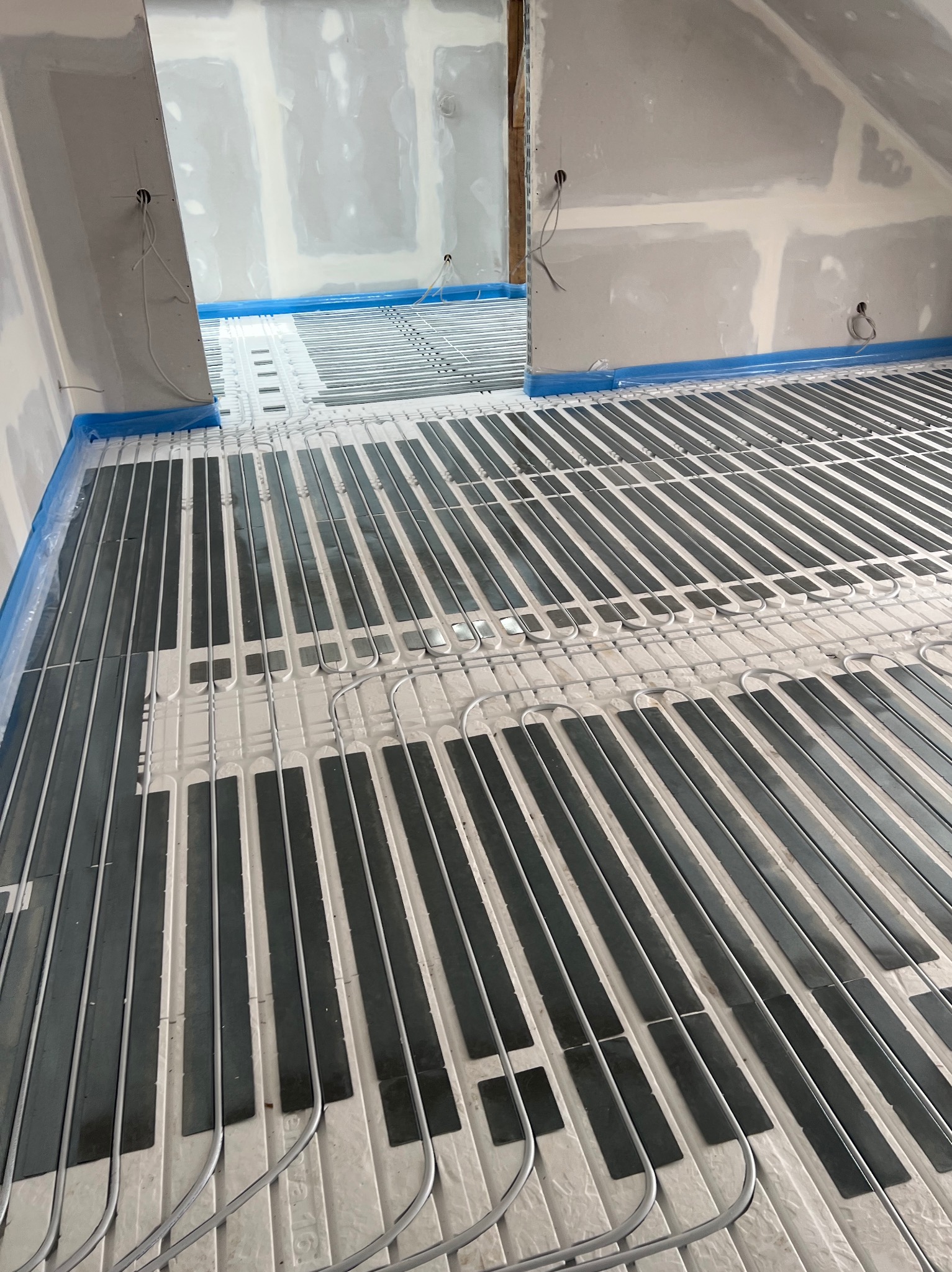 Montaż ogrzewania podłogowego w poddaszu: białe płyty izolacyjne z czarnymi elementami grzewczymi i szarymi rurkami, zabezpieczone niebieską folią przy ścianach.