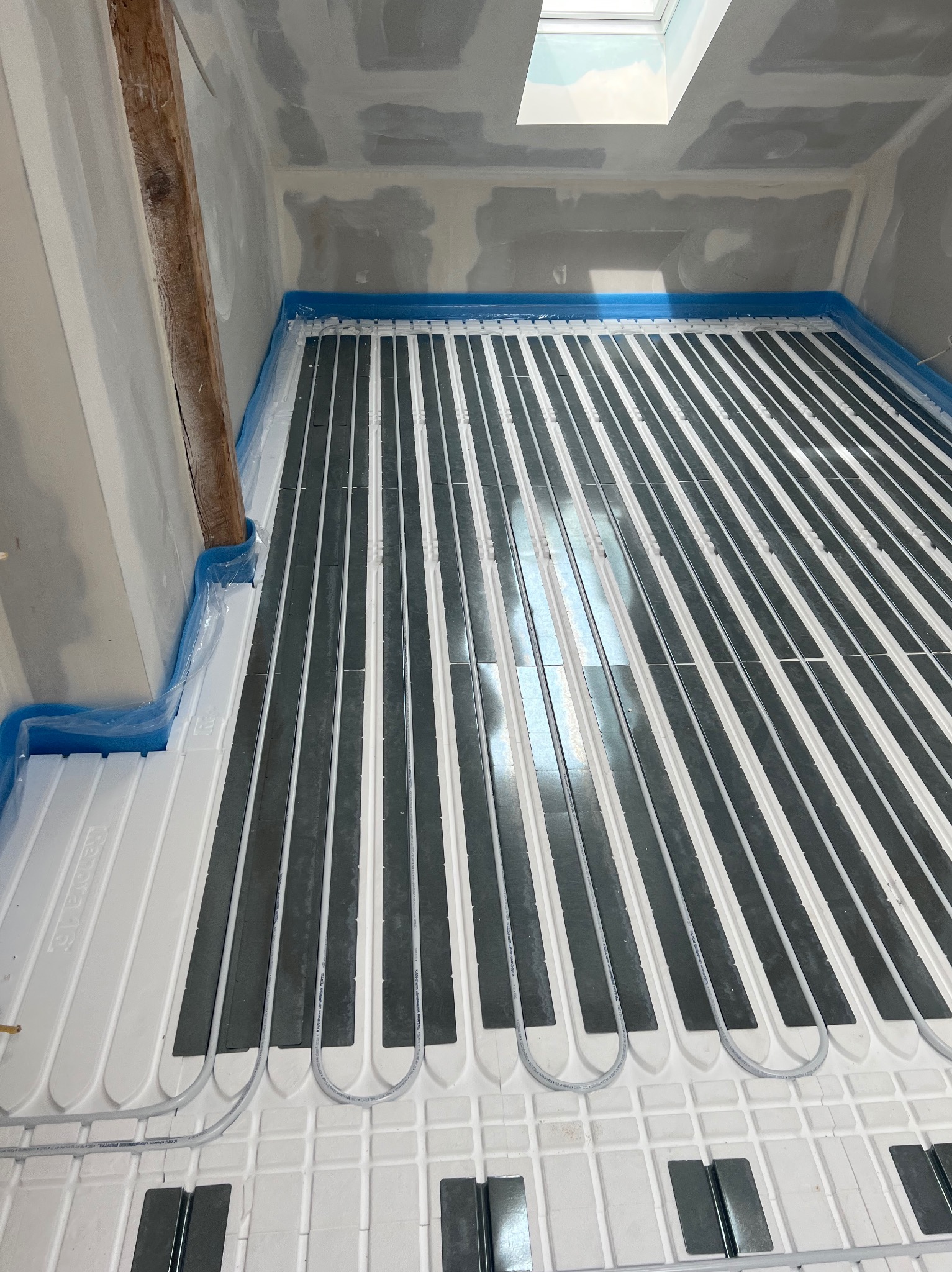 Ułożona instalacja ogrzewania podłogowego na białych płytach izolacyjnych, zabezpieczona folią przy ścianach, widoczny fragment drewnianej belki stropowej i okno dachowe.