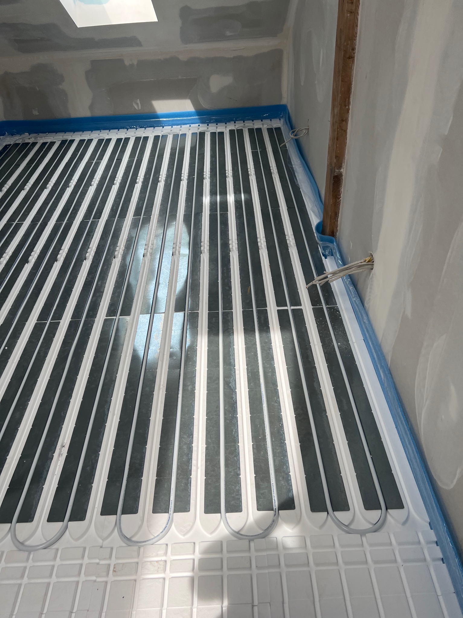 Ułożona instalacja ogrzewania podłogowego na białej macie, z widocznymi szarymi płytkami grzewczymi i rurami, zabezpieczona folią przy ścianach w surowym stanie.