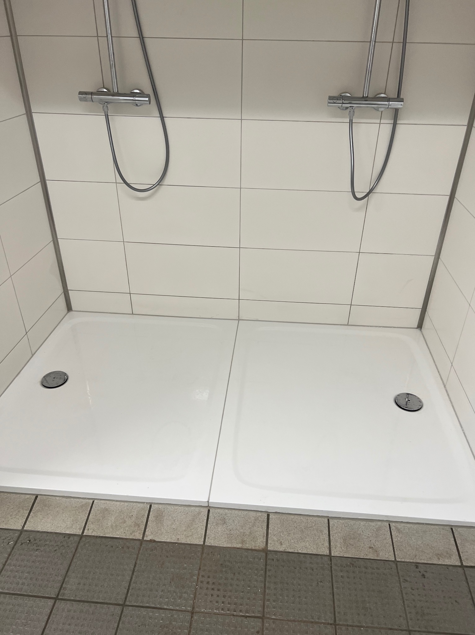 Dwie białe kabiny prysznicowe z chromowanymi bateriami i odpływami, wyłożone białymi płytkami, z szarą podłogą. Widok z góry, symetryczny układ.